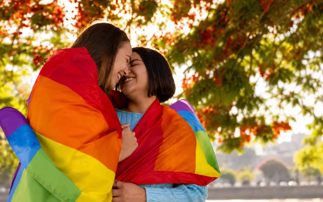 Two LGBTQ+ individuals wrapped in a rainbow Pride flag sharing a joyful and supportive moment outdoors.